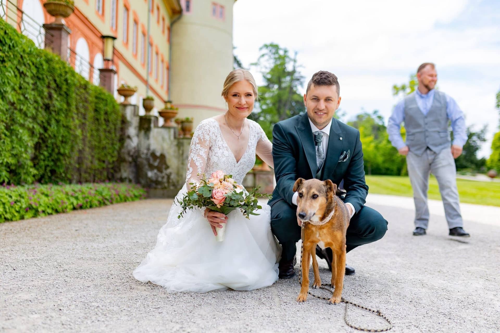 Verena Marius Hochzeitsfotograf Fotograf Worms Mettenheim 08.SFDK4Sdq Brautpaar posiert zusammen mit ihrem braunen Hund auf dem Kiesweg vor dem gelb-orangen Schloss Herrnsheim