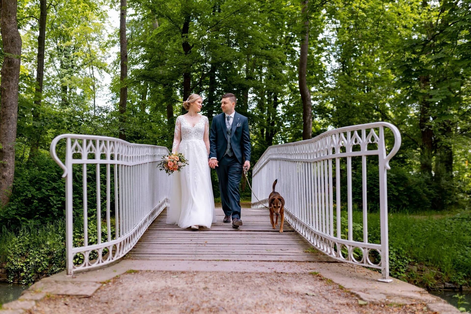 Verena Marius Hochzeitsfotograf Fotograf Worms Mettenheim 09.D479CAwP Brautpaar und Hund spazieren entspannt über die weiße Bogenbrücke durch den grünen Schlossgarten