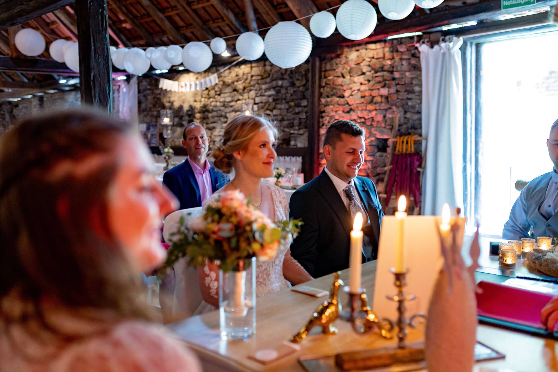 Verena Marius Hochzeitsfotograf Fotograf Worms Mettenheim 17.DBPWt46f Brautpaar sitzt am langen Holztisch mit Gästen im rustikal dekorierten Festsaal mit weißen Laternen und warmem Kerzenlicht