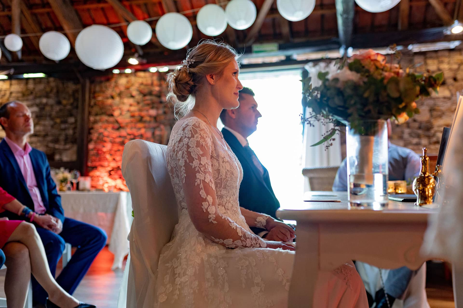 Verena Marius Hochzeitsfotograf Fotograf Worms Mettenheim 19.Pgg3hxP Blonde Braut sitzt an weißem Tisch unter hängenden weißen Laternen in historischem Steingemäuer